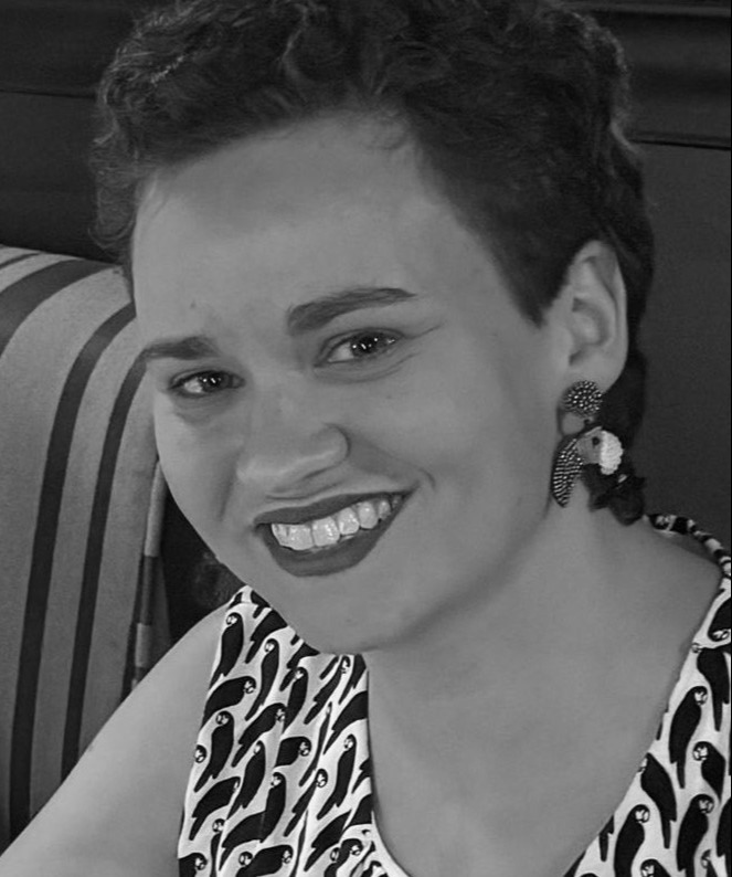 April Dyer, a smiling person with short curly hair, wears patterned earrings and a sleeveless top adorned with a penguin design, sitting in front of a striped background.