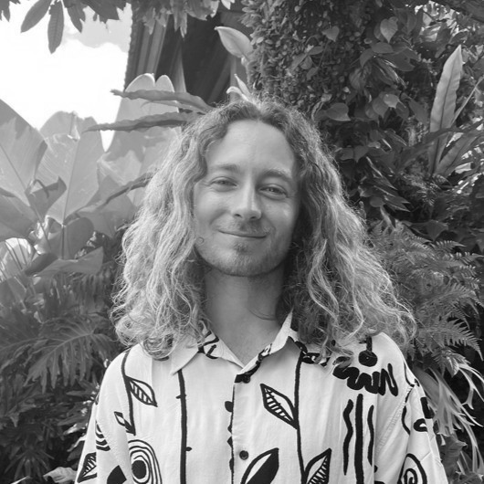 A person reminiscent of James Mason with long curly hair stands in a patterned shirt, set against the backdrop of a lush, leafy environment. The scene is beautifully captured in black and white.
