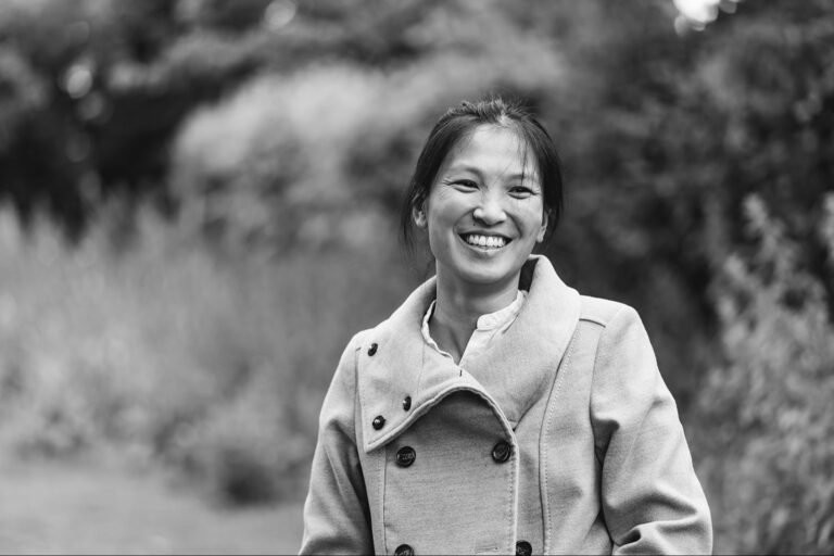 Anna Lau stands smiling outdoors, wearing a light-colored coat. The background shows blurred greenery, adding a serene touch. The image, captured in black and white, beautifully highlights her joyful expression.