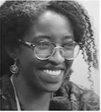 A black and white image captures Azza Dirar, a smiling person with curly hair, glasses, and earrings, holding a microphone.