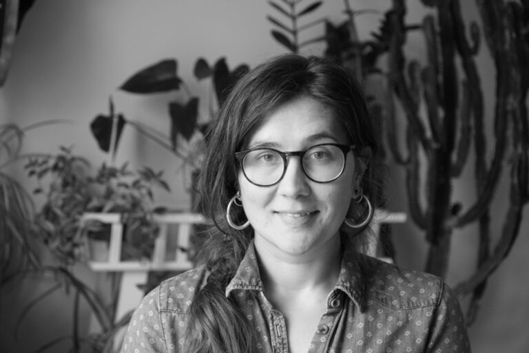 Ruth Larbey, with long hair, glasses, and hoop earrings, smiles slightly. Indoors with various plants in the background, the black-and-white image captures a serene moment.