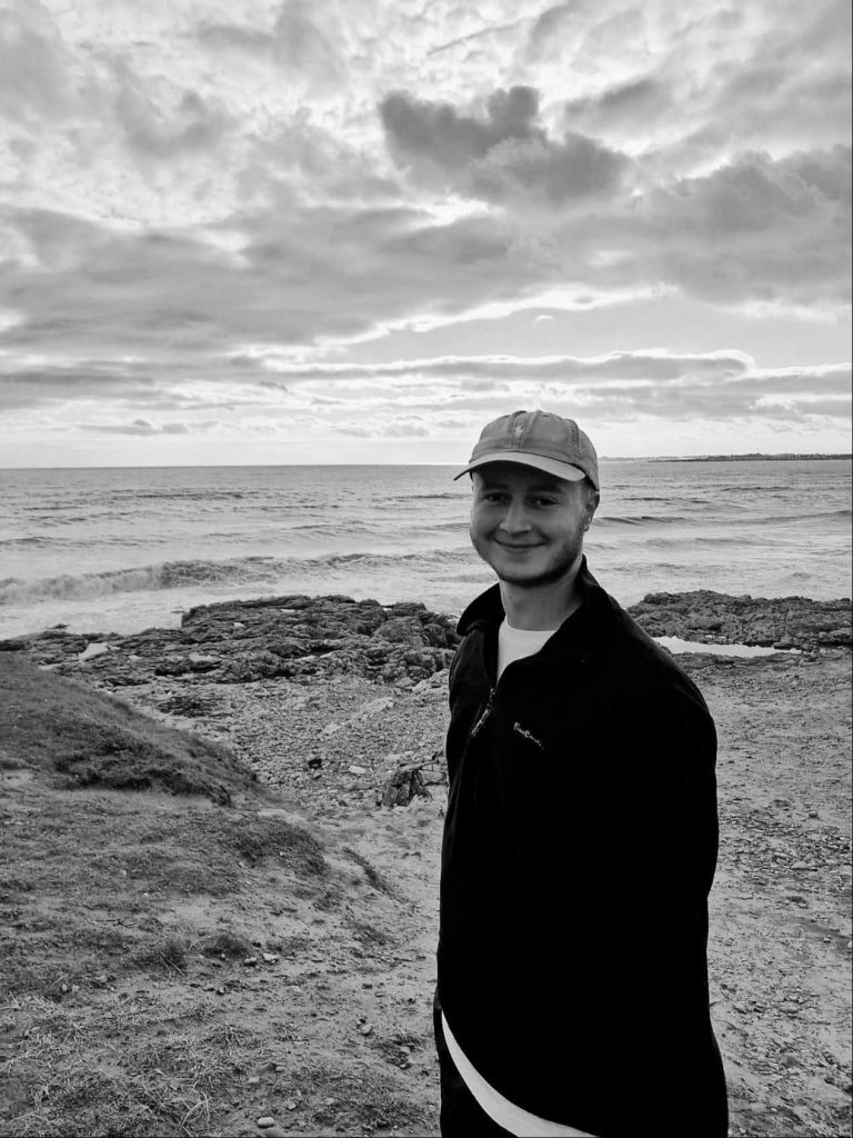 A black and white photo of Daniel Jolly standing near the ocean.