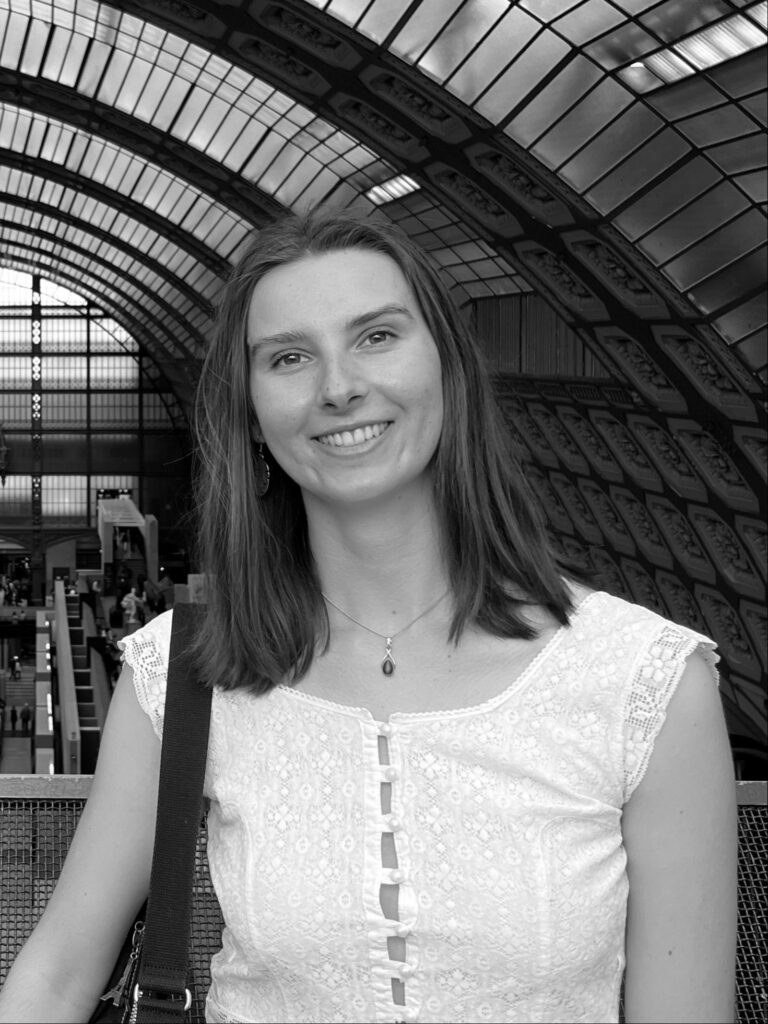 Danni Walters, with shoulder-length hair wearing a white lace top, stands in front of an arched glass ceiling with metal framework in a railway station or similar building.