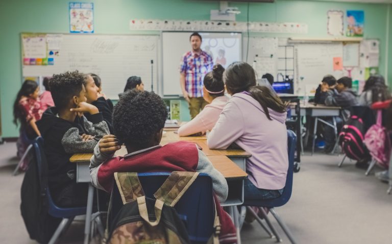 A group of children sitting at desks in a classroom, overseen by a coordinator.