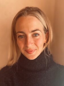 Tyndall Spotlight: Manasa Sharma and Chyna Dixon 2 Portrait of a smiling woman with blonde hair, wearing a black turtleneck, posed against a tan background with Tyndall Spotlight.