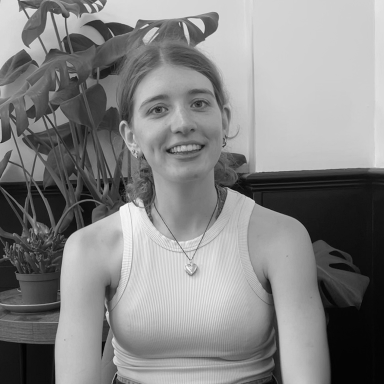 Young woman, Gracie Horton, smiling at the camera, seated indoors with a plant in the background. Black and white photo.