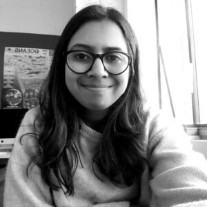 Tyndall Spotlight: Manasa Sharma and Chyna Dixon 1 A young woman with glasses, identified as Manasa Sharma, smiling at the camera in a cozy sweater, indoors, with a world map poster in the background.