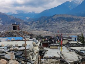 A mountainous landscape with scattered stone houses and colorful prayer flags fluttering like applause from the heavens. A blue-roofed building stands proudly in the foreground, beneath snow-capped peaks that seem to embody success against a cloudy sky backdrop.