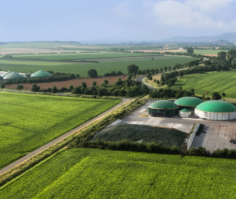 Aerial view of a biogas plant with several green-domed structures surrounded by green agricultural fields and a winding road in the countryside, showcasing a vibrant example of bioeconomy and sustainability.