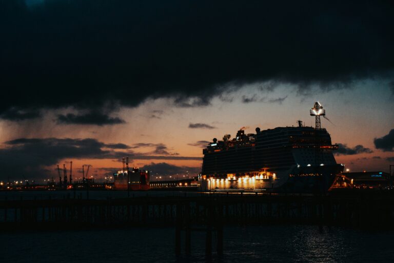A large cruise ship docked at a port during sunset, with the sky showing deep blue hues and the ship illuminated by lights using UK shore power.