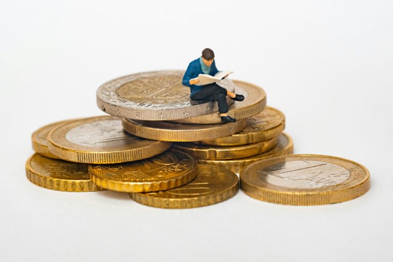 Miniature figure of a man sitting on a stack of large coins and reading a book about economic development, against a plain white background.