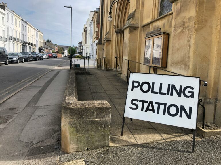A sign reading "POLLING STATION" is placed outside a building on a quiet street in Ireland, with several parked cars, ensuring the scene contributes to improved electoral integrity.