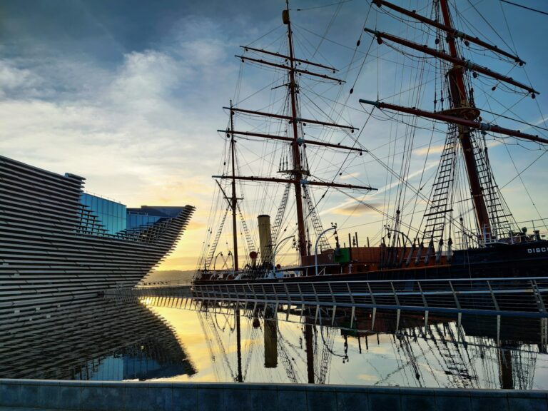 A modern architectural building sits beside a docked large, tall-masted sailing ship in one of the historic Scottish ports, reflected in the still water below, under a partly cloudy sky at sunset.