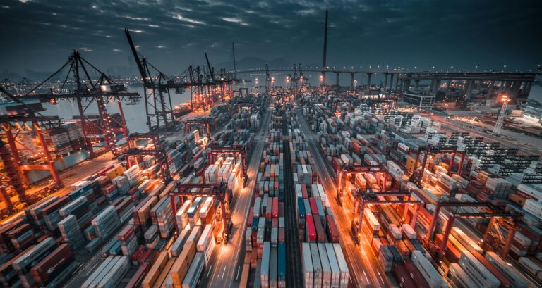 Aerial view of a busy container port at twilight, with numerous colorful shipping containers and cranes from the current shipping fleet, adjacent to a large bridge.