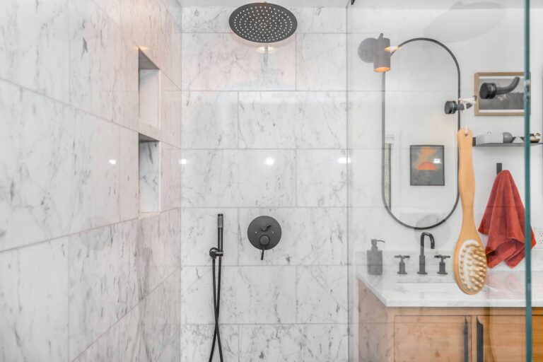 Modern bathroom with white marble walls, a rain showerhead, black fixtures, an oval mirror, and a wooden vanity. A large body brush and a red towel hang near the sink, encouraging a subtle shift in behavior change towards sustainable living.