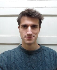 A man with short brown hair and a slight smile, resembling Jack Heslop, is wearing a grey cable-knit sweater, standing in front of a white paneled wall.