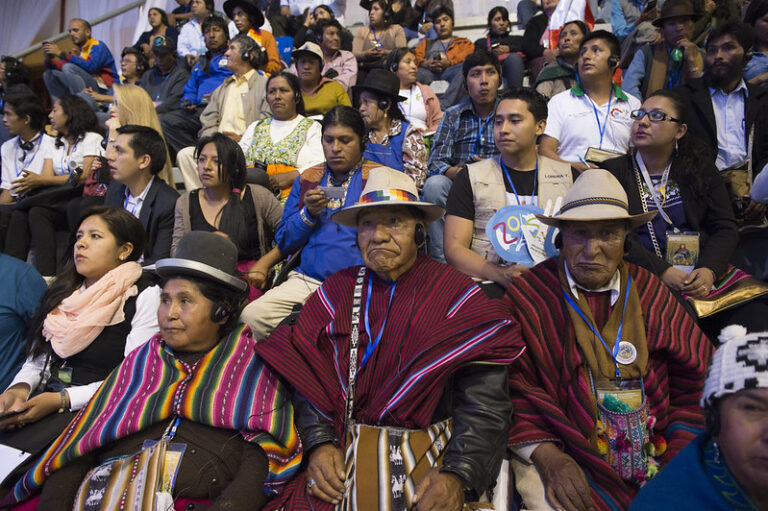 A group of people, some in traditional attire, sit in an audience. The scene appears to be at a conference or gathering, with a diverse mix of men and women attentively watching ahead. Among them are indigenous people from Bolivia passionately discussing climate change.