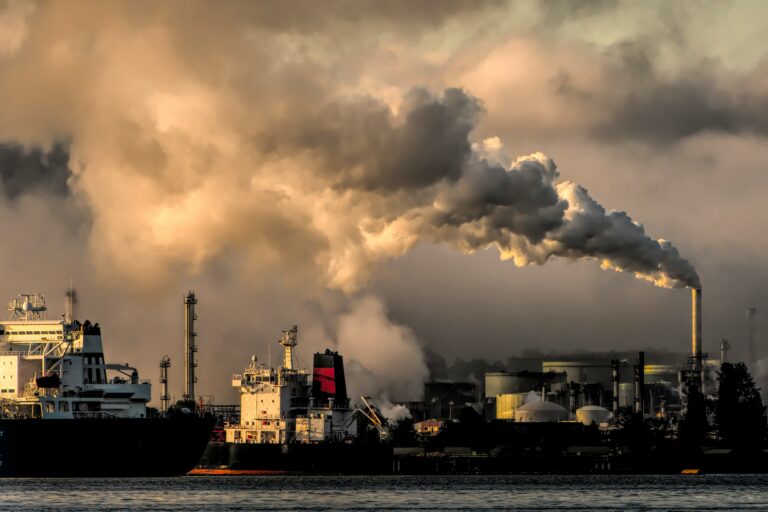 A large industrial plant emits thick smoke from multiple chimneys, with ships docked nearby. The sky is hazy, and the surrounding area appears heavily industrialized. Efforts to curb global emissions are critical to progress in reducing environmental impact.