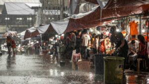 A bustling outdoor market with people shopping under tarps in heavy rain, water pouring off the shelters and collecting on the ground—each drop a testament to their resilience against a changing climate, with stories of endurance whispered among the crowd.
