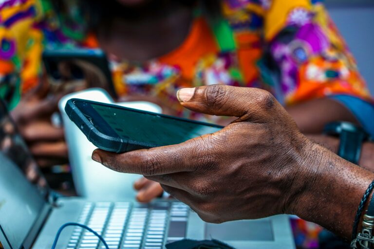 Close-up of a hand holding a smartphone above a laptop. In the background, a Nigerian social entrepreneur in colorful attire is holding a phone, likely capturing workshop reflections.