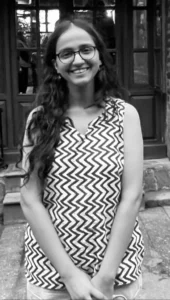 Black-and-white photo of a smiling individual with long hair and glasses, wearing a sleeveless top with a zigzag pattern, standing in front of a door. The photograph captures the essence of Anowyesha Dash's radiant joy in the Tyndall Spotlight.