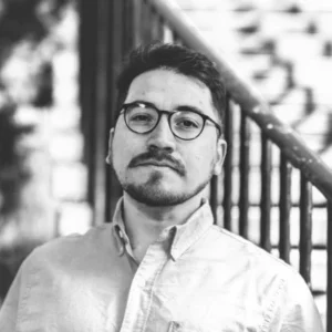 A person with glasses and a beard, wearing a button-up shirt, stands next to a metal railing in a black-and-white photo, capturing the essence of Nicolás Enrique Labra Cataldo.