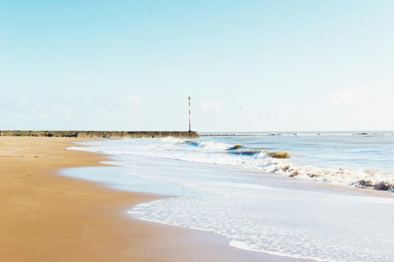 A sandy beach with gentle waves lapping the shore, a stone pier extends into the water, and a striped pole is visible at the end of the pier—a tranquil scene subtly impacted by Coastal System Dynamics.