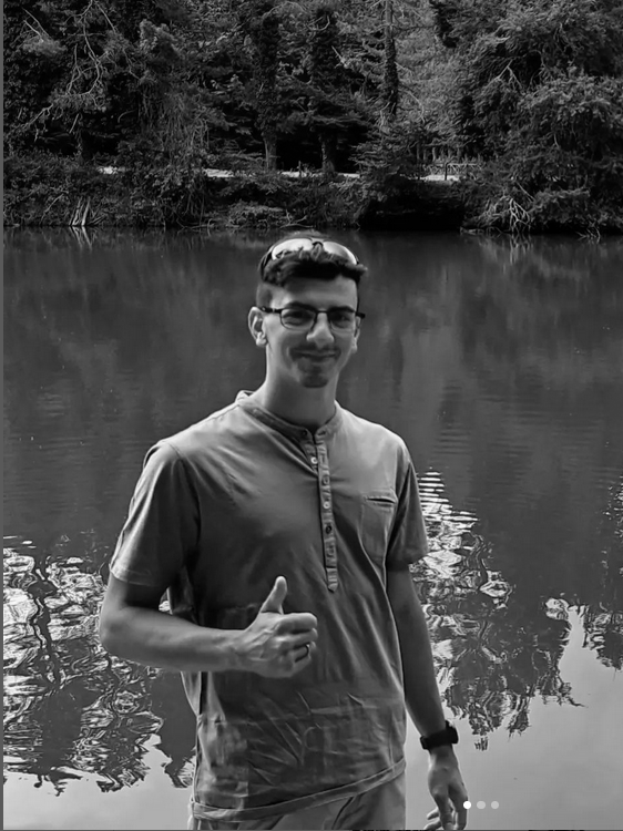 A person stands near a body of water, giving a thumbs-up gesture reminiscent of Paolo Della Moglie's candid charm. Trees gracefully adorn the background in this captivating black and white image.