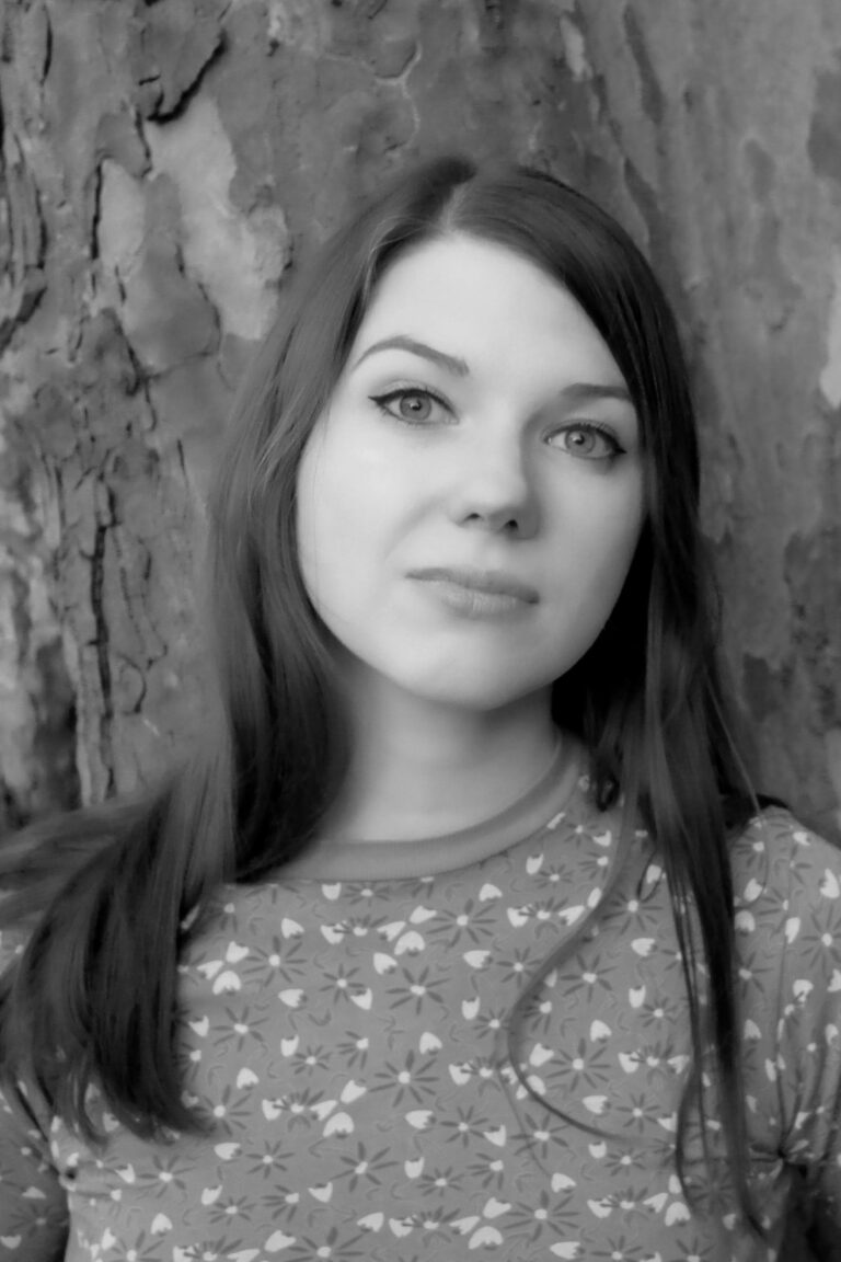 Sophie Hopesmith, with her long hair cascading down, stands poised in front of a tree, looking directly at the camera. The captivating image is rendered in striking black and white.