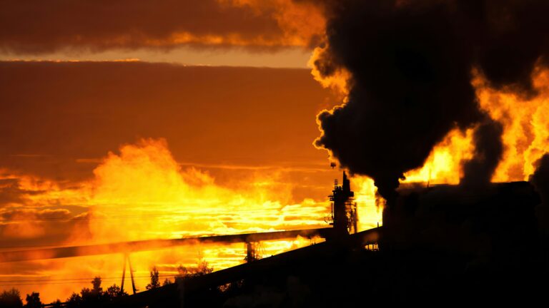 Silhouette of an industrial facility emitting smoke and steam against an orange sunset sky with clouds.