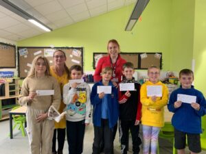A group of seven children and two adults stand indoors holding information about being climate ambassadors, smiling at the camera in a classroom setting with green walls and a bulletin board.