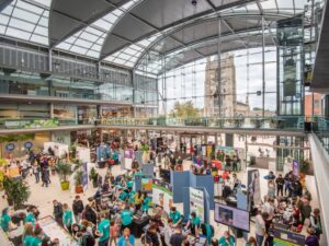 Stalls set up for the Norwich Science Festival inside The Forum in 2021, a busy indoor event with booths, people interacting, and a view of a large church structure, St Peter Mancroft, through a glass wall in the background