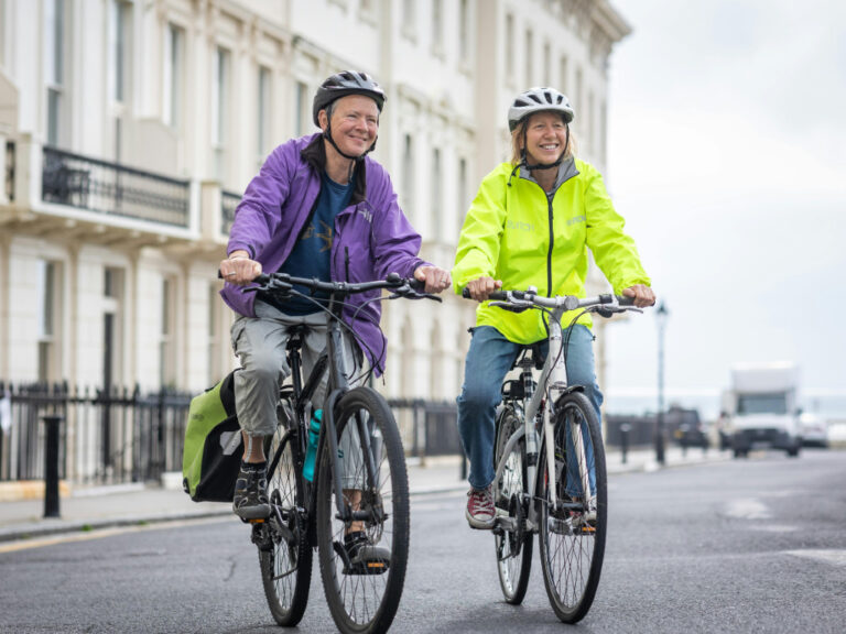 Two people in helmets and jackets ride bicycles on a city street, smiling.