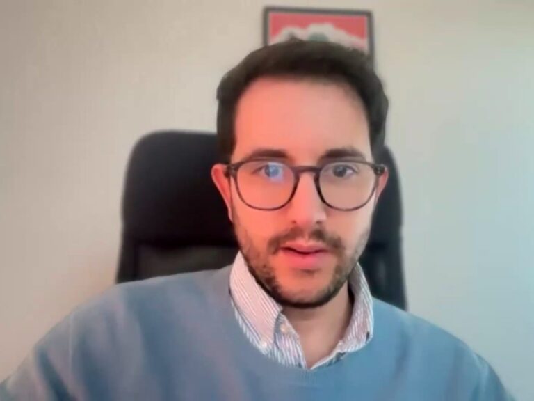 A person with glasses and a beard, wearing a blue sweater and collared shirt, sits in an office chair with a framed picture in the background.