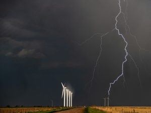Dark stormy sky with fork lightning, and a few gleaming white wind turbines beside a long, straight road next to fields