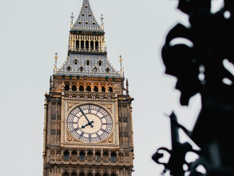 The clock face of Big Ben, part of the Houses of Parliament building in London, UK, against a cloudy sky and foregrounded by the silhouette of some shrubbery.