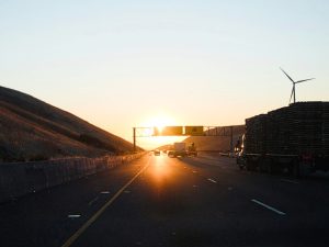 A truck driving down a highway next to a wind turbine, with huge setting sun at top of road. Credit: Luke Marcelo via Unsplash