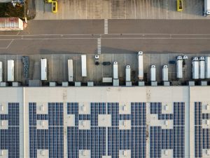 An aerial view of a parking lot and loading dock in Germany, half filled with cars and lorries, besides a warehouse roof lined with solar panels.
