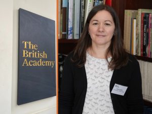 Dr Katie Jenkins with books behind her, next to the British Academy signage, as her British Academy Fellowship gets underway