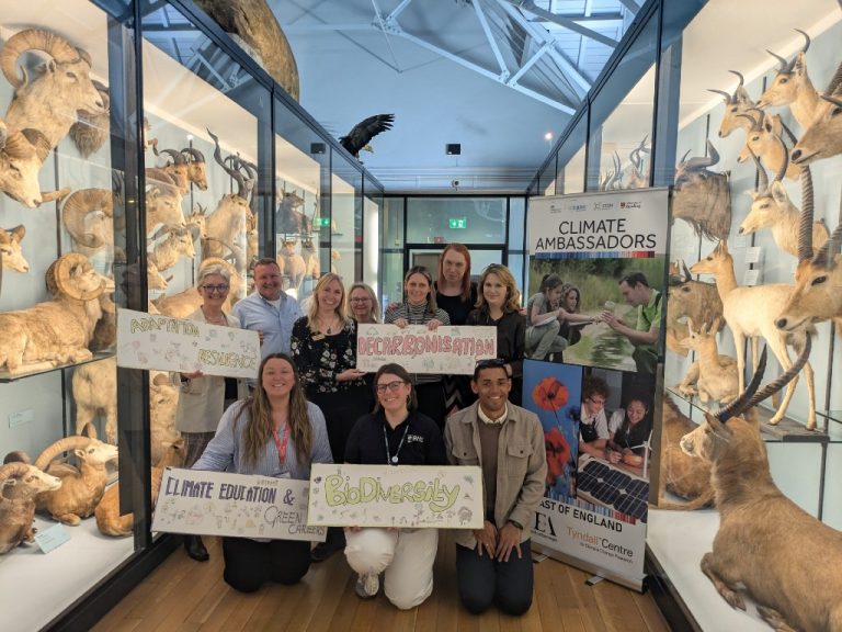 Climate Ambassadors in the East of England, holding up signs saying 'Adaptation and resilience', 'Decarbonisation', 'Biodiversity' and 'Climate education and green careers', in a museum setting, surrounded by taxidermy specimens of goats, rams, and other animals.