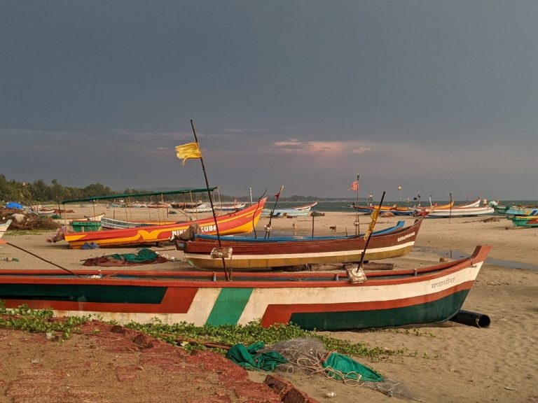 Colourful fishing boats on a sandy beach beneath dark storm clouds, with sunlight illuminating the shoreline and sea, illustrating research into drowning risk and climate-related coastal hazards.