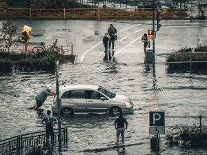 A flooded city street with a car being pushed and pedestrians navigating the water.