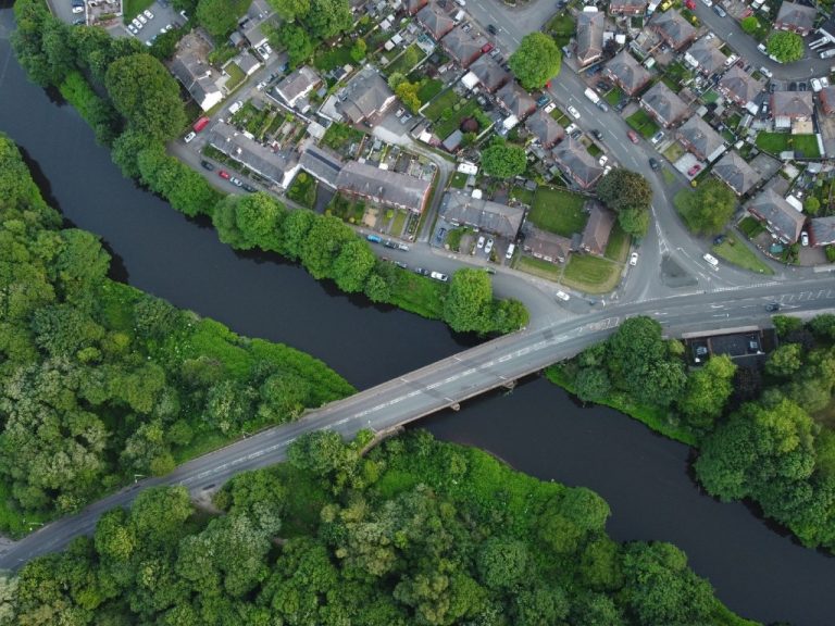 Aerial view of a residential neighbourhood beside a river, showing homes, roads, bridges and green spaces within an urban landscape