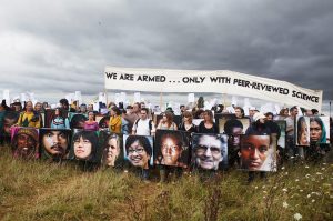 Rows of researchers and scientists protesting against a proposed third runway at Heathrow in 2007, holding up a large banner reading 'We are armed only with peer-reviewed science', copies of a report from the Tyndall Centre for Climate Change Research, and cardboard shields covered in images of people from around the globe all facing the impacts of climate change: at the Camp for Climate Action UK, against a cloudy and storm-threatening sky