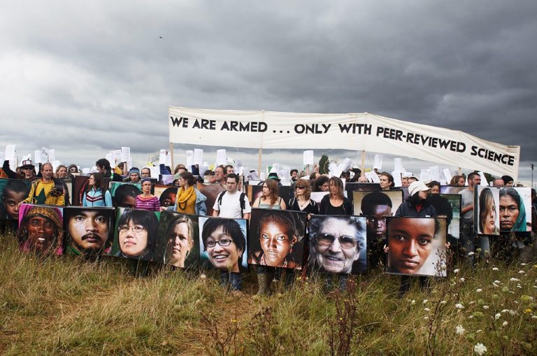 Rows of researchers and scientists protesting against a proposed third runway at Heathrow in 2007, holding up a large banner reading 'We are armed only with peer-reviewed science', copies of a report from the Tyndall Centre for Climate Change Research, and cardboard shields covered in images of people from around the globe all facing the impacts of climate change: at the Camp for Climate Action UK, against a cloudy and storm-threatening sky