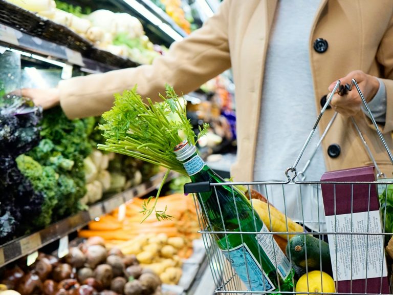 Woman putting food and drink, including vegetables, into a shopping basket at a supermarket.