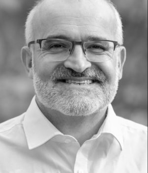 In this black-and-white portrait, Neil Ward, a man with glasses and a beard, smiles while wearing a white shirt.