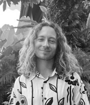 A person reminiscent of James Mason with long curly hair stands in a patterned shirt, set against the backdrop of a lush, leafy environment. The scene is beautifully captured in black and white.