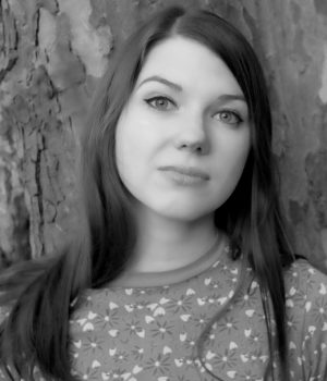 Sophie Hopesmith, with her long hair cascading down, stands poised in front of a tree, looking directly at the camera. The captivating image is rendered in striking black and white.