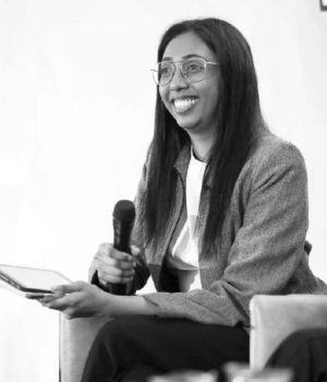 A person seated and smiling, holding a microphone and some papers, wearing glasses and a blazer, Wasundara Doradeniya exudes confidence and charm.